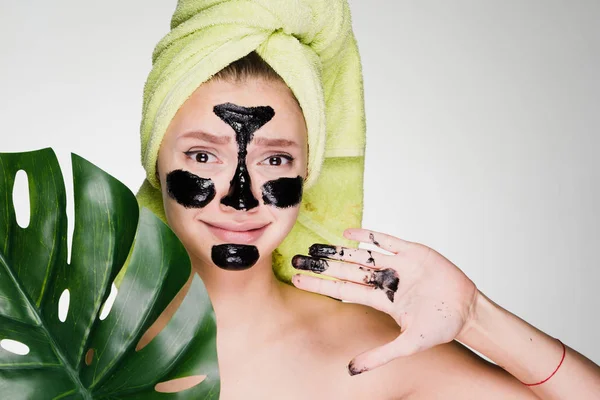 Funny young girl with a green towel on her head puts on her face a black mask against acne — Stock Photo, Image