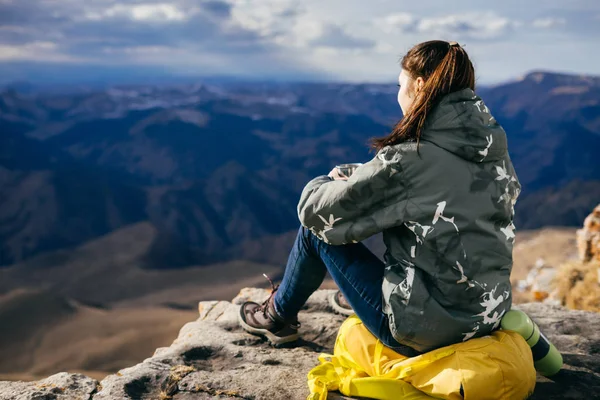 Активна молода дівчина сидить на краю скелі на жовтому рюкзаку, насолоджується гірською природою — стокове фото