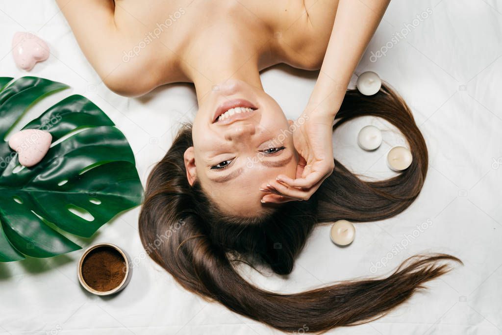 smiling long-haired girl enjoying spa treatments, scented candles and coffee scrub