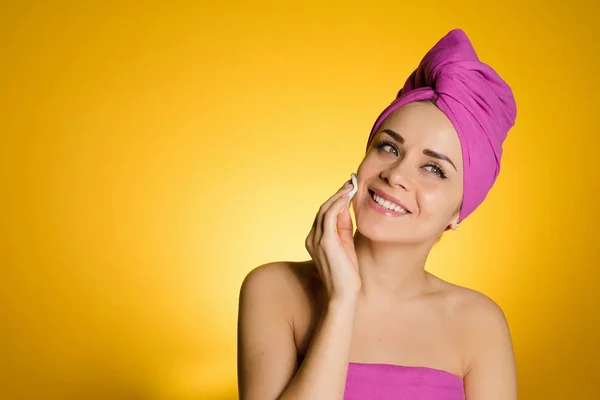 Glückliches junges Mädchen mit rosa Handtuch auf dem Kopf reinigt Gesicht mit Baumwollkissen — Stockfoto