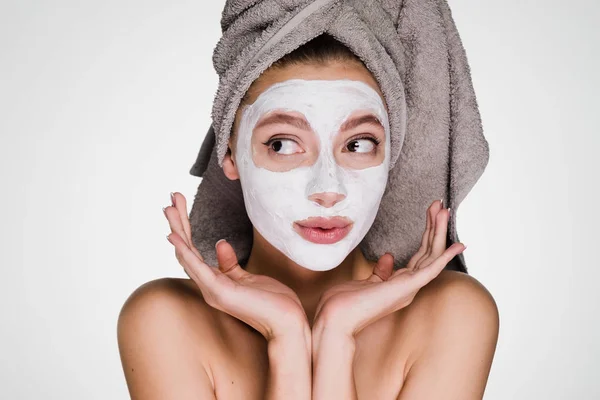 Funny young girl with a towel on her head applied a white moisturizing mask to her face — Stock Photo, Image