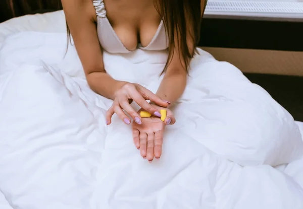 Young girl sits in bed under a white blanket, holds yellow earplugs against street noise — Stock Photo, Image