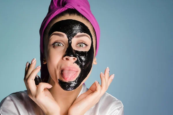Engraçada jovem com uma toalha rosa na cabeça colocou uma máscara preta em seu rosto, mostra sua língua — Fotografia de Stock