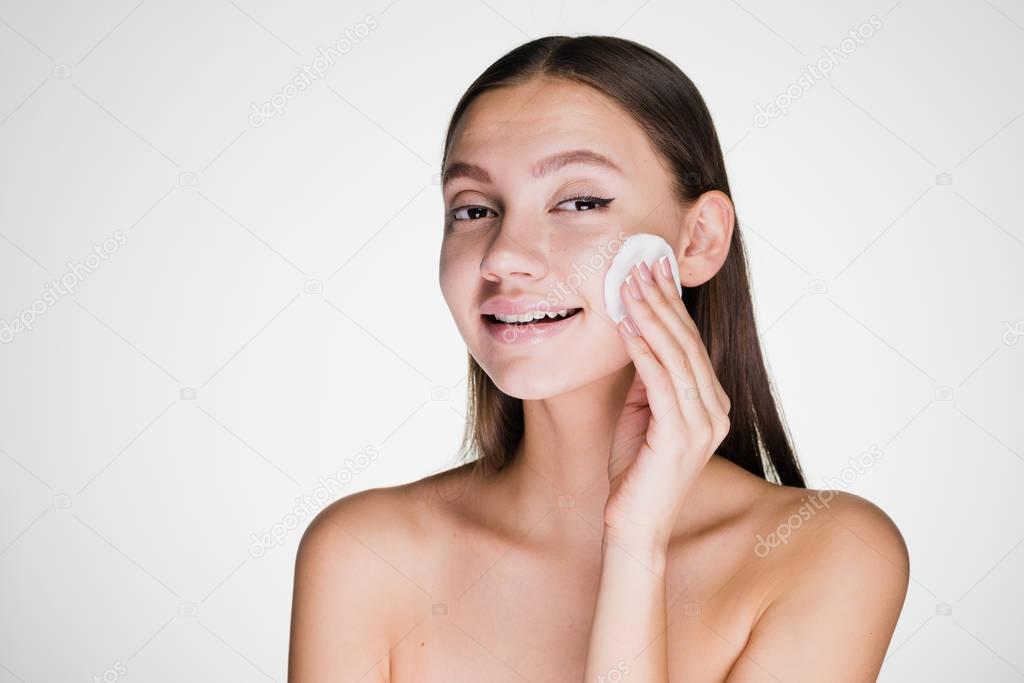 cute smiling girl cleanses the skin on her face with a cotton ball