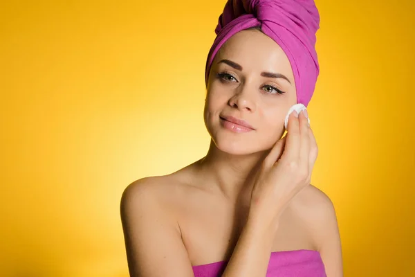Mulher com uma toalha na cabeça em um fundo amarelo remove maquiagem com um disco de algodão — Fotografia de Stock