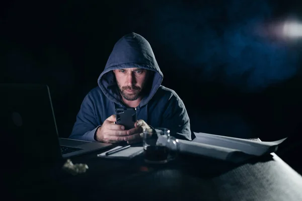 Τύπος χάκερ με ένα σακάκι με κουκούλα κάθεται στο laptop και κοιτάζοντας το τηλέφωνο — Φωτογραφία Αρχείου