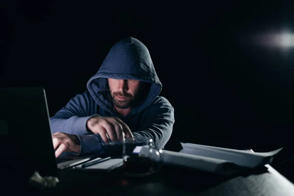 Guy hacker with a jacket with a hood sits behind a laptop — Stock Photo, Image