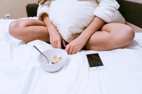 On the bed a woman sits next to the phone and a plate of oatmeal porridge — Stock Photo, Image