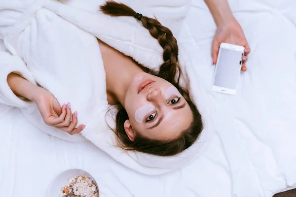Vrouw ligt op bed met witte vlekken op haar gezicht bedrijf een telefoon — Stockfoto