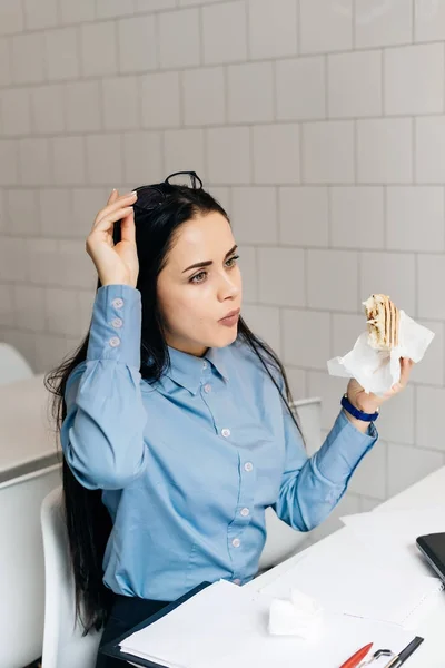 Женщина сидит в офисе за столом и ест сэндвич — стоковое фото