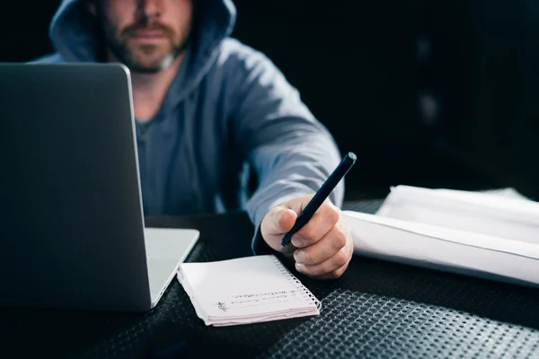 a man hacker sits behind a laptop and writes something to a notebook