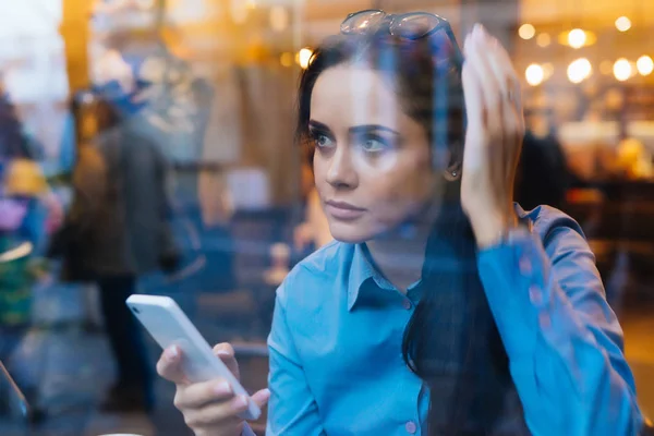 Young busy girl in a blue shirt is sitting in a cafe, is holding a smartphone — Stock Photo, Image
