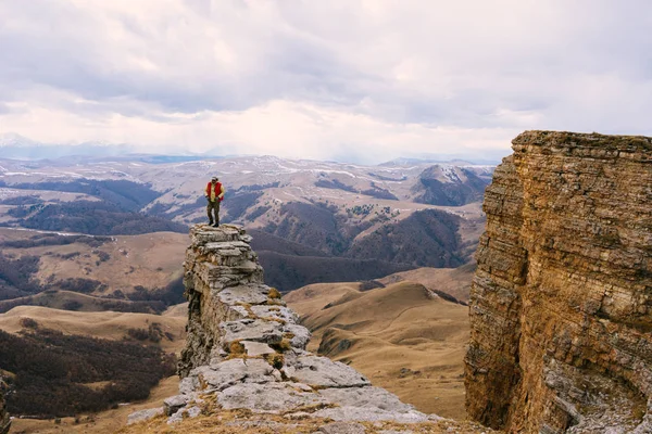 Мужчина путешественник в красной куртке стоит на краю скалы и наслаждается горной природой — стоковое фото