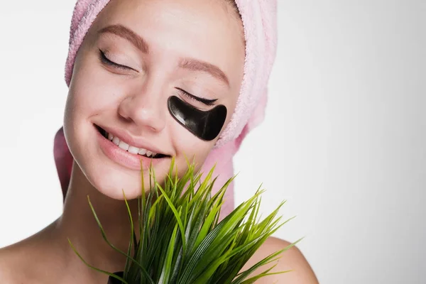 Ευτυχισμένος χαμογελαστό κορίτσι με μια ροζ πετσέτα στο κεφάλι της, κάτω από τα μάτια του μαύρα μπαλώματα, κατέχει ένα πράσινο φυτό — Φωτογραφία Αρχείου