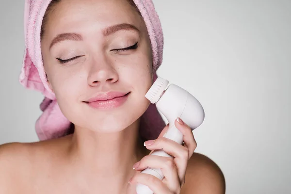 Happy smiling girl with pink towel on head doing deep cleansing of facial skin with electric brush, eyes closed — Stock Photo, Image