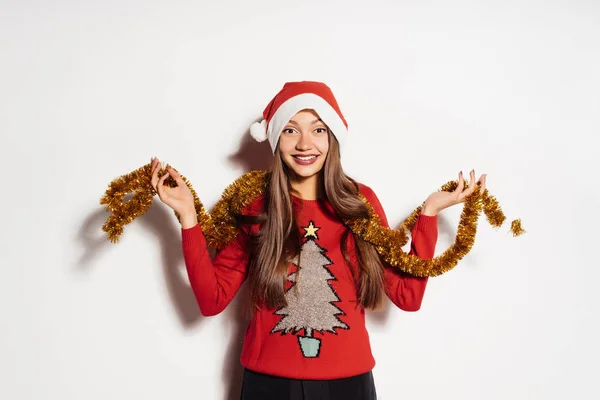 Menina feliz em um boné vermelho como Papai Noel segurando um ouropel de ouro, esperando o ano novo — Fotografia de Stock
