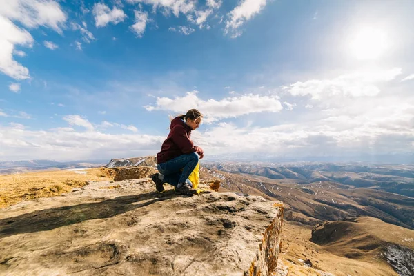 Active young girl sits at the edge of the mountain, enjoys nature and the sun, holds a yellow backpack — Stock Photo, Image