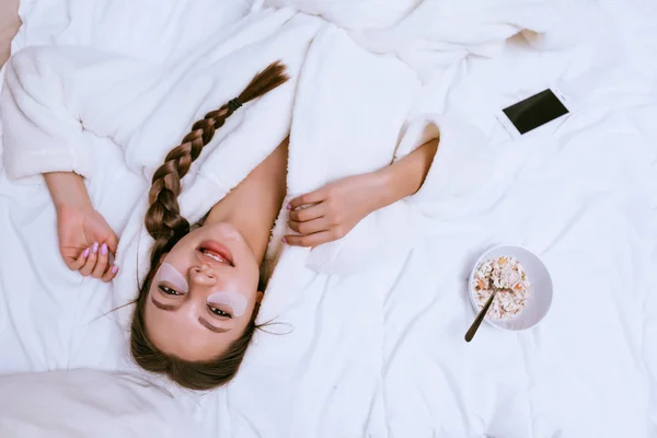 A cute young girl in a white robe lies on a bed, under the eyes of a patch, next to a plate of oatmeal porridge — Stock Photo, Image