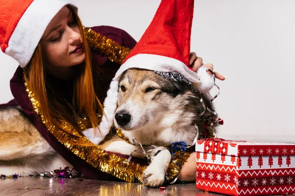 Happy red-haired girl in a red cap like Santa Claus sits on the floor with her dog, New Year 's atmosphere, golden tinsel and gifts — стоковое фото