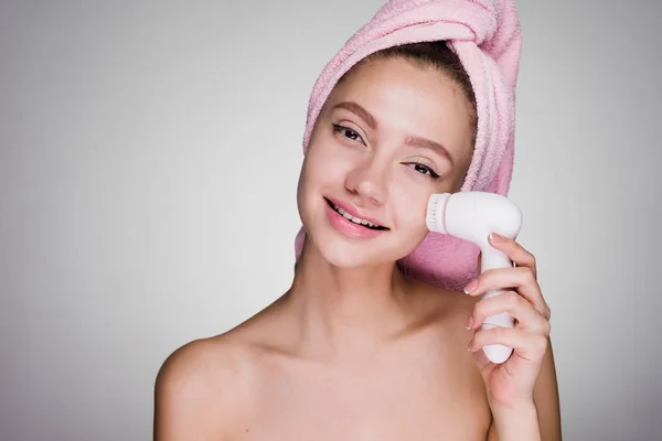 Bella ragazza con un asciugamano rosa sulla testa fa una pulizia profonda della pelle del viso con l'aiuto di una spazzola elettrica — Foto Stock