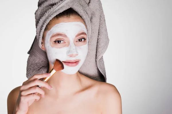 Joven chica atractiva con una toalla n agolove pone una máscara blanca nutritiva en la cara con un cepillo —  Fotos de Stock