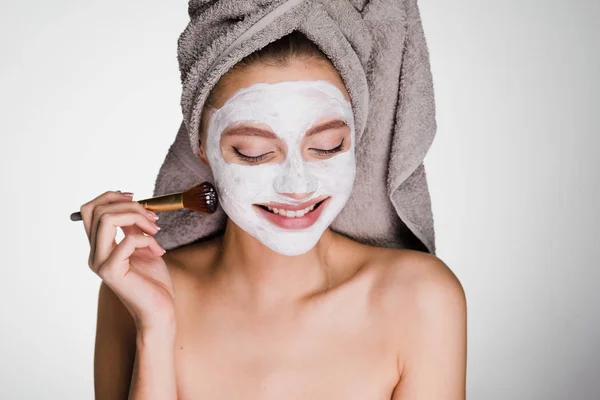 Šťastná dívka s ručníkem na hlavě použití bílé hydratační maska na obličej s kartáčem — Stock fotografie