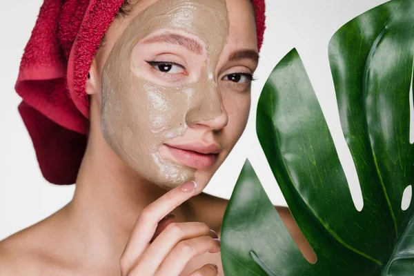 Una giovane ragazza attraente con un asciugamano rosso sulla testa applicato una maschera di argilla a metà del viso, una procedura spa — Foto Stock