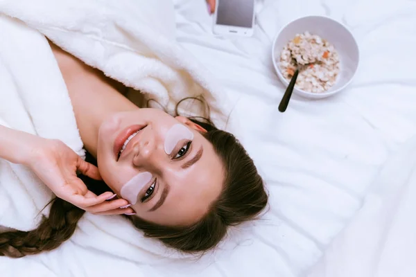 Linda chica sonriente se encuentra en la cama, bajo los ojos manchas blancas, junto a un plato de avena gachas —  Fotos de Stock