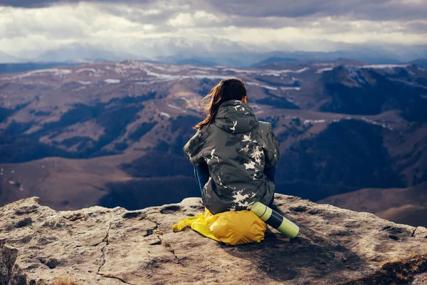 Молода дівчина з жовтим рюкзаком сидить на краю скелі, насолоджуючись гірською природою — стокове фото