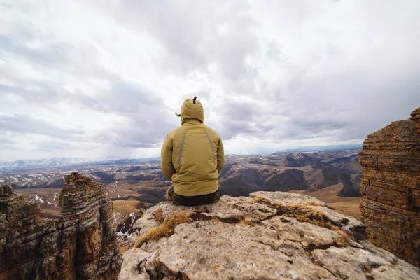 Un viajero masculino en una chaqueta se sienta en el borde de un acantilado y disfruta de la naturaleza de la montaña —  Fotos de Stock