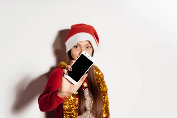 Junges Mädchen mit roter Mütze auf dem Kopf zeigt Handybildschirm, Weihnachtsstimmung — Stockfoto
