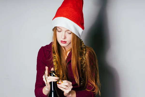Молодий руда дівчина з Червону Шапочку як Санта-Клаус на голову відкриває пляшку з шампанським — стокове фото