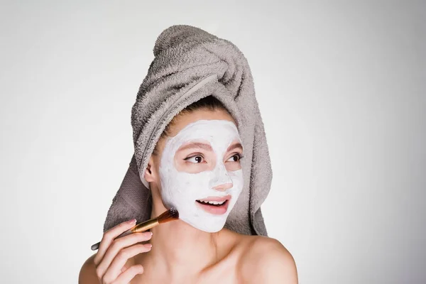 Linda chica sonriente con una toalla en la cabeza aplicando una máscara blanca nutritiva en su cara con un cepillo —  Fotos de Stock