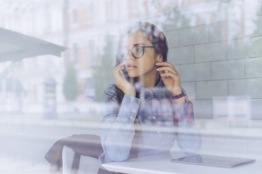 genç kız mavi gömlek ve gözlük bir kafede oturmuş telefonda konuşuyor