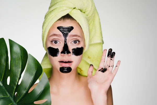 A surprised young girl with a green towel on her head applied a black mask to the problem areas on her face, her hands were soiled — Stock Photo, Image