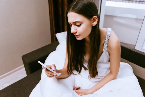 Niña somnolienta se sienta en la cama debajo de una manta blanca, mira en su teléfono inteligente —  Fotos de Stock