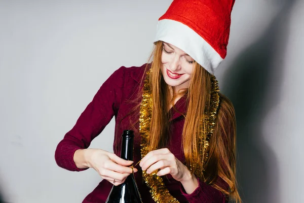 Щаслива руда дівчина з червоною новорічною шапочкою на голові відкриває пляшку шампанського, святкує Різдво — стокове фото