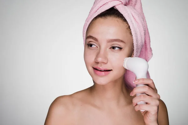 Bella ragazza con un asciugamano rosa sulla testa facendo una pulizia profonda del suo viso con una spazzola elettrica, sorridente — Foto Stock