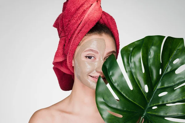 Egy aranyos fiatal lány, egy piros törülközőt a fején alkalmazott agyagos maszk fél arc — Stock Fotó