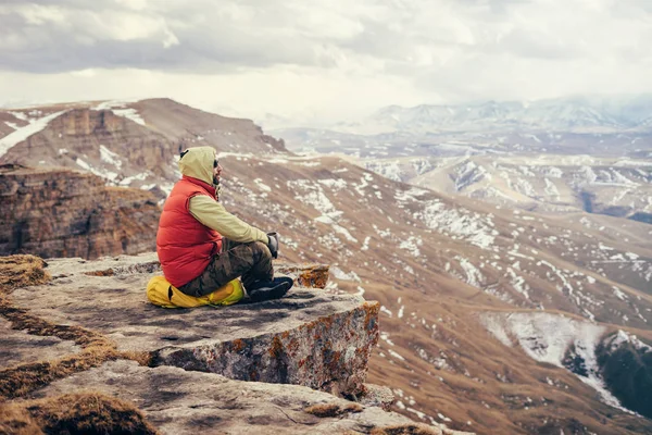 Male traveler in a red jacket sits on the edge of the mountain, on a yellow backpack, enjoying nature — Stock Photo, Image