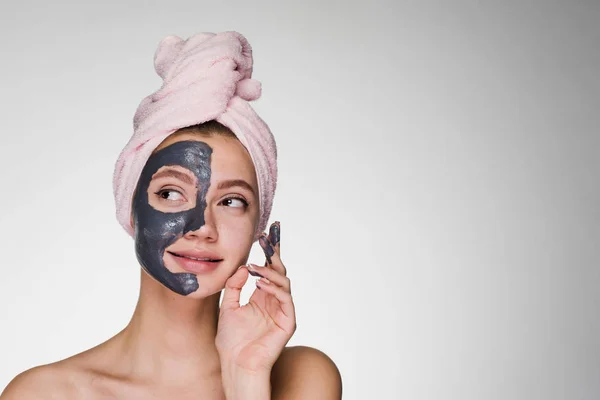 Ein nettes Mädchen mit einem rosa Handtuch auf dem Kopf hat eine Tonmaske auf ihr halbes Gesicht aufgetragen, schaut verträumt weg — Stockfoto