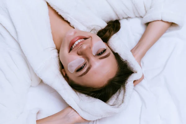 Heureuse fille souriante en robe de chambre éponge blanche couchée dans le lit, sous les yeux patchs — Photo