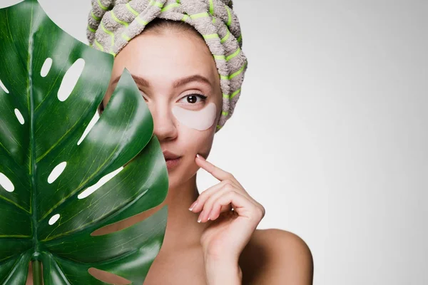 Cute young girl with a towel on her head, holds a green leaf, under the eyes patches, enjoys a spa — Stock Photo, Image
