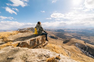 aktif genç kız sarı bir sırt çantası ile bir uçurumun kenarında oturur, doğa ve güneş sahiptir