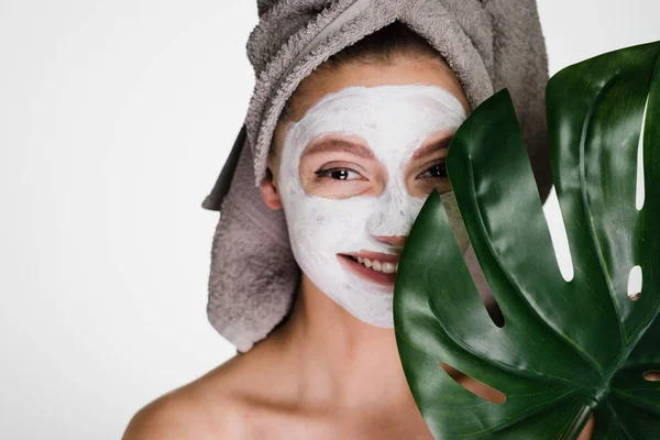 Ένα γέλιο κορίτσι με μια πετσέτα στο κεφάλι της εφαρμόζεται μια λευκή ενυδατική μάσκα στο πρόσωπό της, κατέχει ένα πράσινο φύλλο, ημερήσιο σπα — Φωτογραφία Αρχείου