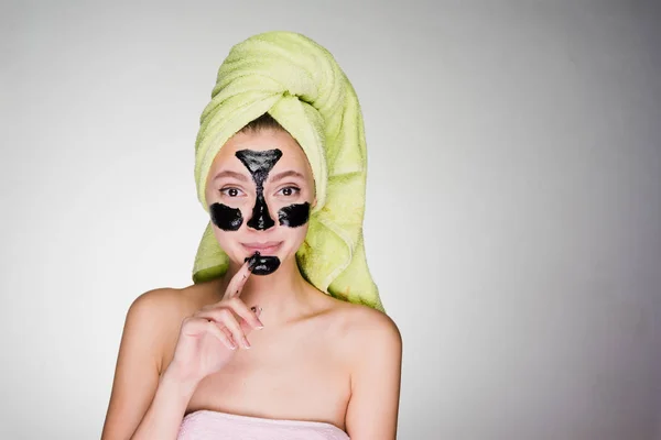 Ένα χαριτωμένο νεαρή κοπέλα με μια πράσινη πετσέτα στο κεφάλι της εφαρμόζεται μια μαύρη μάσκα για τις ζώνες πρόβλημα στο πρόσωπό της — Φωτογραφία Αρχείου