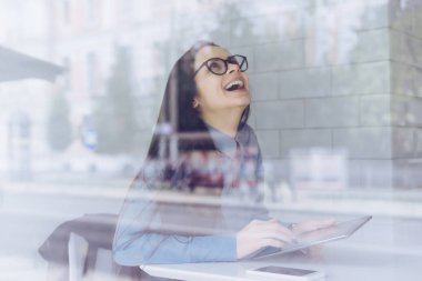 kafede oturan bir tablet tutan gözlük ve mavi gömlekli mutlu gülen kız