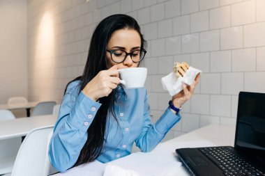 genç kız freelancer dizüstü bilgisayarda çalışan, kafede kahve içme ve sandviç yeme mavi gömlekli