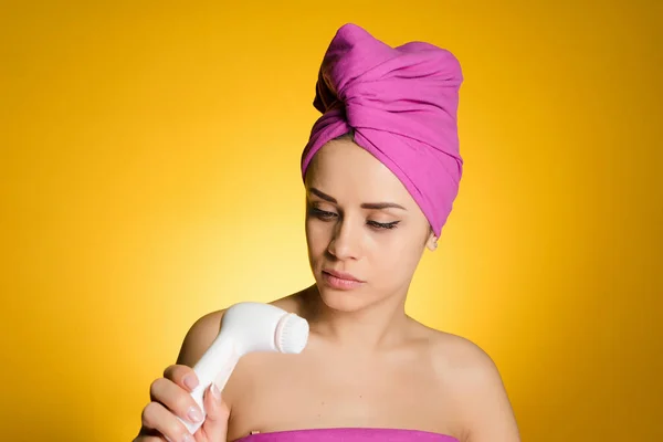 Una giovane ragazza con un asciugamano rosa sulla testa tiene una spazzola elettrica per una pulizia profonda della pelle del suo viso — Foto Stock