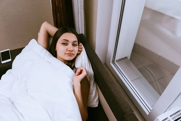 Uma menina sonolenta agradável está na cama sob um cobertor branco, acorda cedo e não quer se levantar — Fotografia de Stock